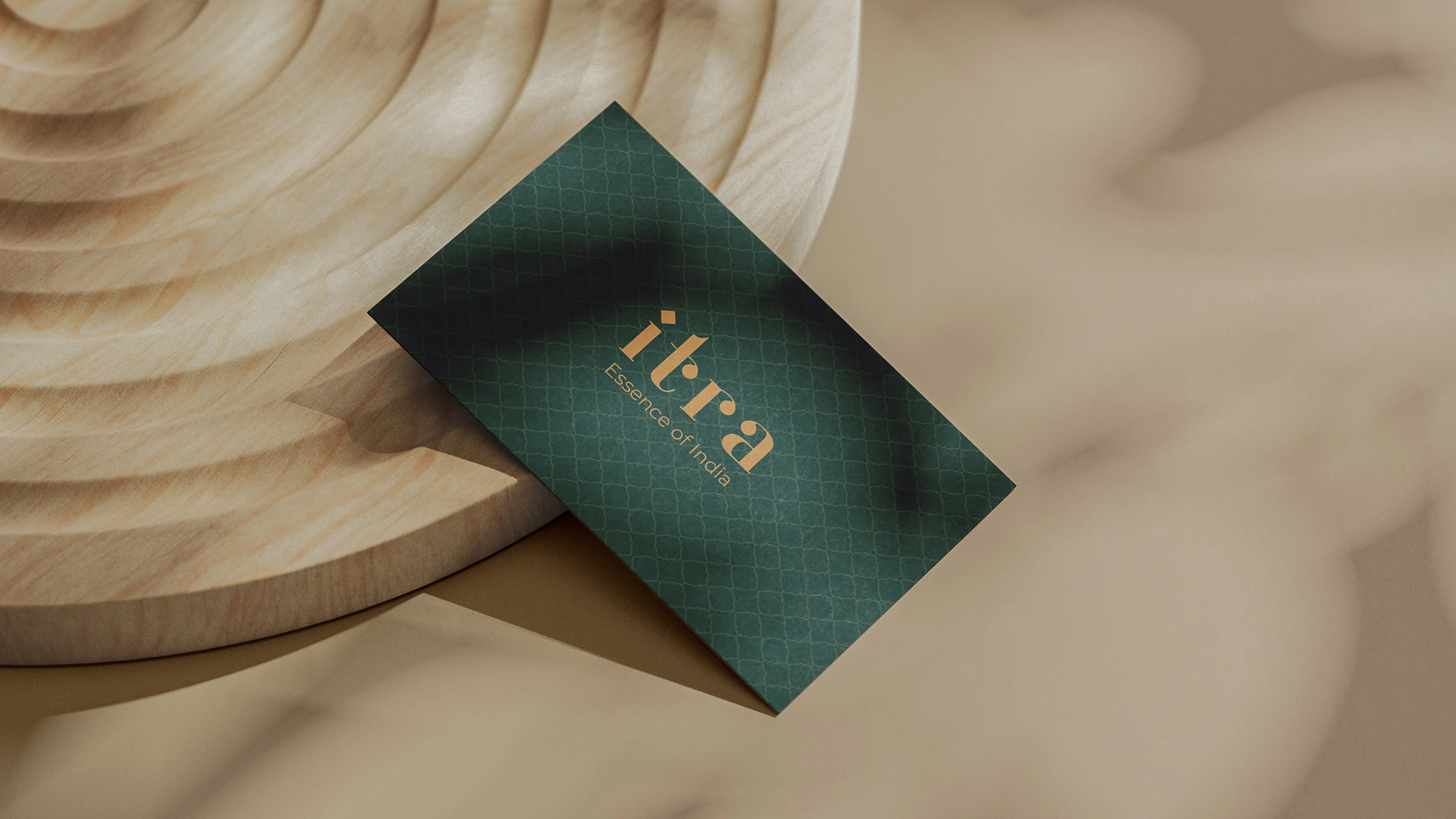 Itra business card in forest green with gold wordmark and quatrefoil lattice pattern, angled on a carved wooden surface with warm natural light